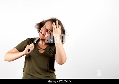 Young brunette woman with tangled unruly hair in casual khaki shirt holding comb and pressing palm to head in pain with closed eyes and open mouth sta Stock Photo
