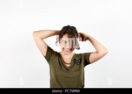 Adorable young tanned female in dark green shirt smiling and looking at camera doing ponytail standing over white studio background Stock Photo