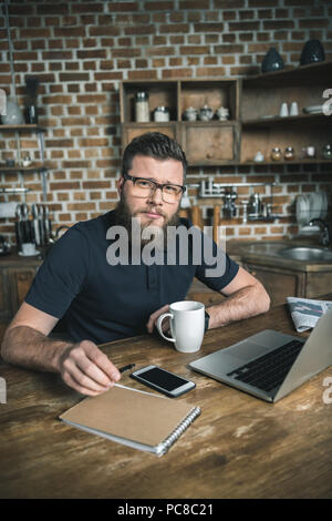 confident freelancer sitting at table with laptop and smartphone while looking at camera Stock Photo