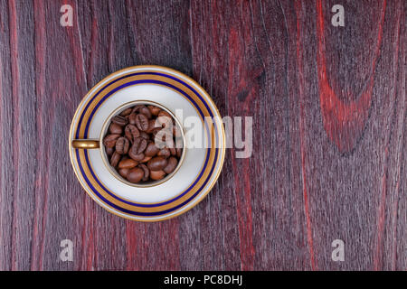 An old cup full of coffee beans with its plate and on top of a mahogany table. Stock Photo
