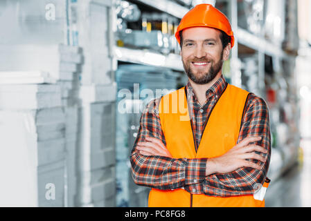 smiling worker in helmet and safety vest with crossed arms in warehouse Stock Photo