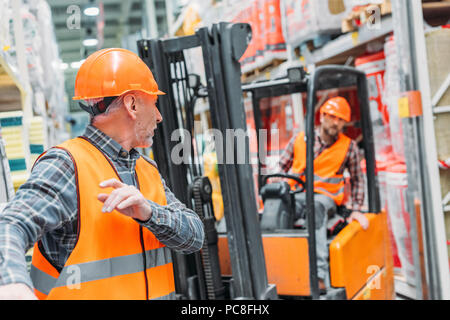 male worker and his colleague working with forklift machine in storehouse Stock Photo