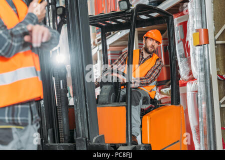 worker in safety vest and helmet sitting in forklift machine in storage Stock Photo