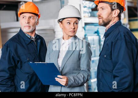 male workers in helmets and female inspector with clipboard in warehouse Stock Photo