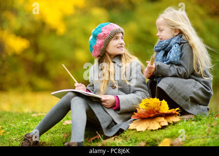 Cute little girls sketching outside on beautiful autumn day. Happy children playing in autumn park. Kids drawing with colourful pencils. Autumn activi Stock Photo