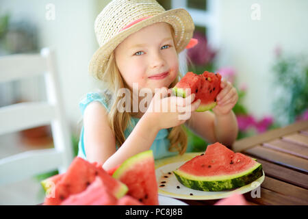 Funny little girl wearing straw hat biting a slice of watermelon outdoors on warm and sunny summer day. Healthy organic food for little kids. Stock Photo