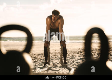 Man doing fitness workout at a beach using battle ropes. Man taking rest during workout with hands resting on knees holding battle ropes and kettlebel Stock Photo