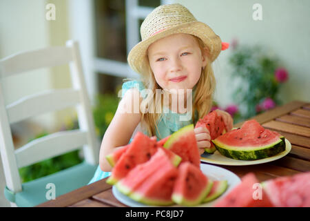 Funny little girl wearing straw hat biting a slice of watermelon outdoors on warm and sunny summer day. Healthy organic food for little kids. Stock Photo