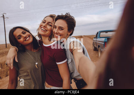 Group of happy young women taking selfie while on road trip. Three young female friends posing for a selfie on country road with a pickup truck in bac Stock Photo