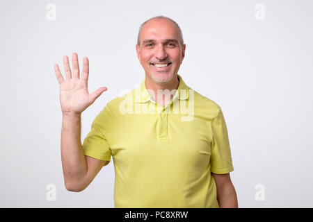 Friendly-looking attractive european man waiving hand in hello gesture while smiling cheerfully. Stock Photo