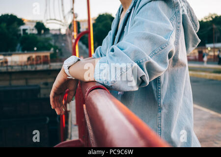 Close-up young woman in jeans jacket standing next to bridge parapet. Candid lifestyle photo of female hands wearing wristwatches leaning on railing c Stock Photo