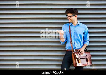 handsome office man with glasses and leather bag, standing against a wall and presenting a side of free space around him Stock Photo