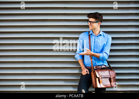 handsome office man with glasses and leather bag, standing against a wall and presenting a side of free space around him Stock Photo