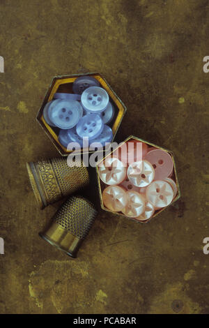 Two small hexagonal brass pill boxes containing blue and pink buttons lying on tarnished brass with two antique brass thimbles Stock Photo