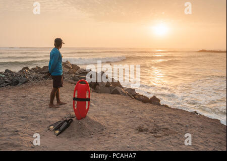 Lifeguard on duty seen from behind, looking over the ocean from a sunset beach. Red float and black flippers on the sand, waves crashing into rocks. Stock Photo
