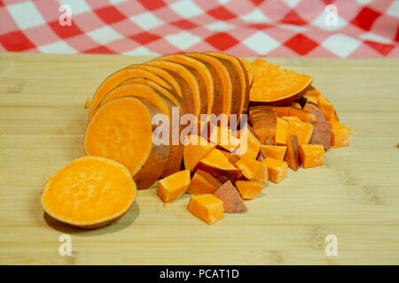 Sliced sweet potato on a cutting board with a red and white checkered table cloth.  Part of the potato has already been diced. Stock Photo