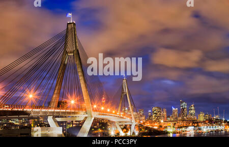 Modern urban bridge in Sydney city CBD against distant towers and cloudy susnet sky with Australian anzac flag on tall column with lights and illumina Stock Photo