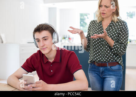 Teenage Boy Wearing Headphones And Using Mobile Phone Being Nagged By Mother At Home Stock Photo