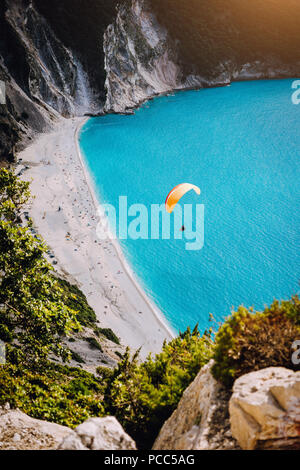 Myrtos Beach, Kefalonia Island, Greece. Figure of a parachutist skydiver with orange parachute against a blue lagoon bay beach. Stock Photo