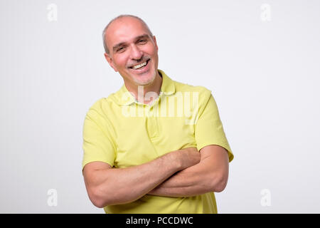 Charming handsome mature man in casual yellow wear keeping arms crossed and smiling Stock Photo