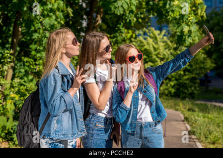 Three girls are teenage girlfriends. In summer after school, in the park. Photographs on a smartphone. Wear jeans clothes and sunglasses. Happy smiling smile they say hello. Stock Photo