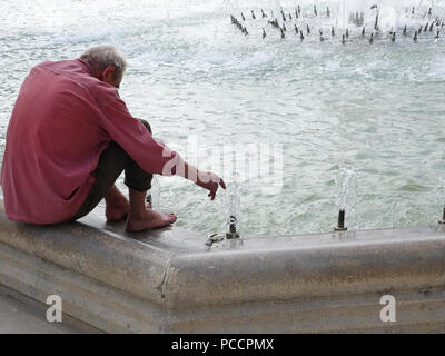 old man feeling hot and uncomfortable in the heatwave tries to keep cool by putting his feet in the cool fountain water Stock Photo