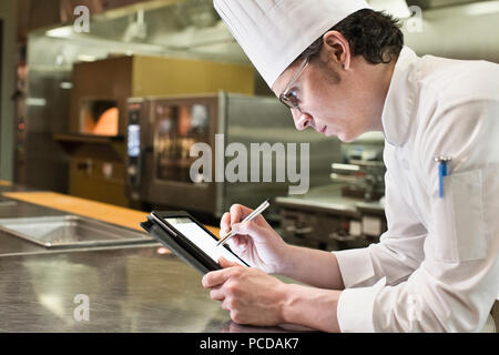 A caucasian male chef working on a notebook computer in a commercial kitchen. Stock Photo