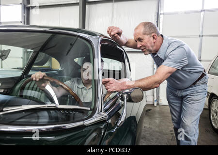 A caucasian senior male car mechanic shows his grandson the driver's view from the driving seat of an old car in a classic car repair shop. Stock Photo