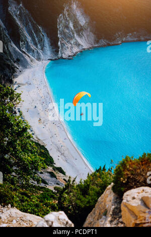 Myrtos Beach, Kefalonia Island, Greece. Figure of a parachutist skydiver with orange parachute against a blue lagoon bay beach. Stock Photo