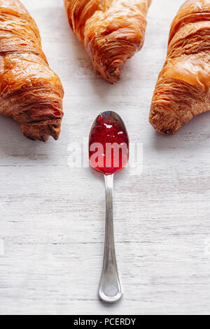 Three croissants on a white wooden table with spoon filled with jam in the middle Stock Photo