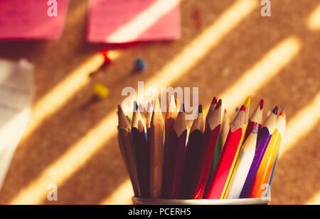 Bunch of colorful pencils in a holder in front of cork board, some push pins and post it, and with rays of sunshine on it Stock Photo