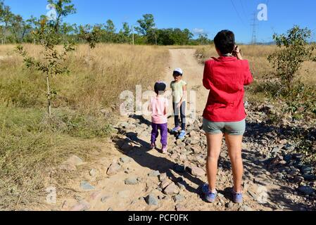 A mother takes a photo of children walking down a dry dusty barren road, Townsville, Queensland, Australia Stock Photo