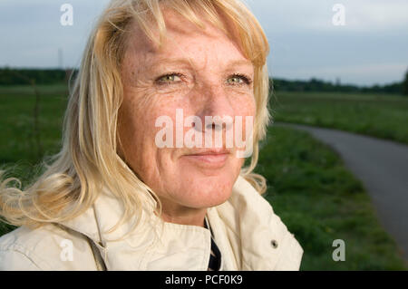 Side head portrait of a middle-aged blond woman in front of green meadow looking cheerfully into the camera. Stock Photo