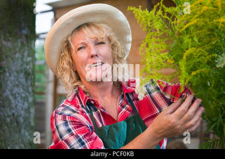 Head-and-shoulder portrait of a blond woman with freckles and straw hat in plaid shirt touching a bunch of flowers in the garden and smiling happily. Stock Photo