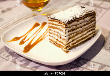 A piece of honey cake with custard and chocolate honey shortcakes on decorated plate and glass of brandy on the background. Stock Photo