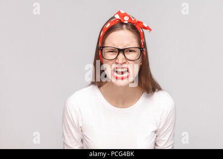 angry face screaming portrait of anger crazy bossy emotional young woman in white t-shirt with freckles, black glasses, red lips and head band. indoor Stock Photo