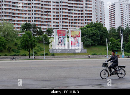 Cyclist passes the inspiration posters that abound in Pyongyang streets across an empty road, North Korea Stock Photo