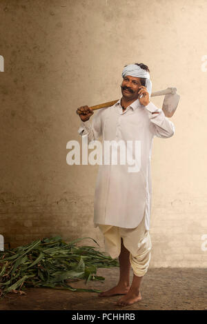 Indian farmer talking on mobile phone while carrying hoe on his shoulder Stock Photo
