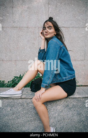 Portrait of fashionable girl sitting near the gray building, strands of hair fluttering by wind over her face. Teen female posing in city dressed in s Stock Photo