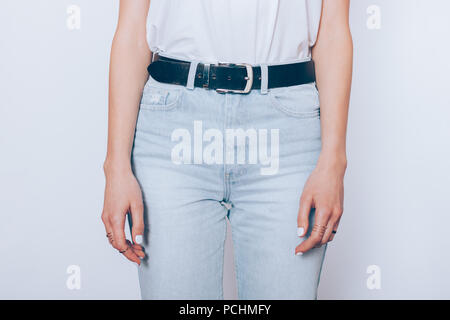 Slender young woman wearing blue high waist mom's jeans with belt and plain t-shirt standing over white background, close-up. Stock Photo