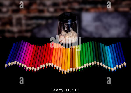 A row of colored pencils lined up in a wavy pattern with a blurry background and an electirc pencil sharpener full of shavings, all set on a black tab Stock Photo