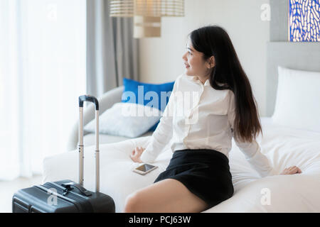 Smiling Asian business woman sitting on bed in hotel room. Business travel concept.