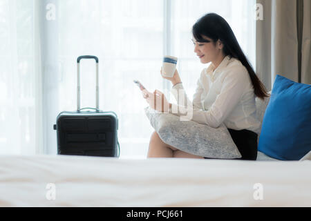 Beautiful Asian young smiling businesswoman in suit drinking coffee and using mobile phone while sitting on the sofa in hotel room. Business travel. Stock Photo