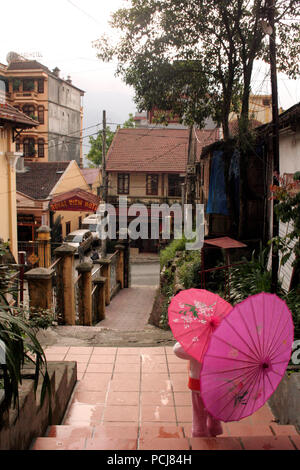 Young Northern Vietnamese girl child twoholding two parasols looking into the distance in a small town at the top of some tiled stairs Stock Photo