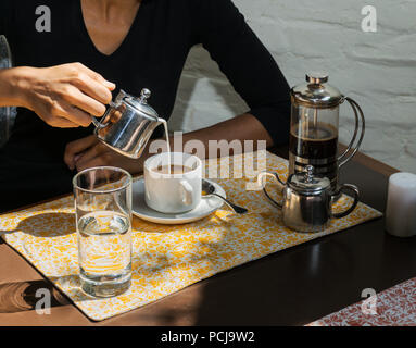 A woman's hand is pouring fresh milk from a pot into a coffee cup, with a French press and metal service, in a restaurant. Stock Photo