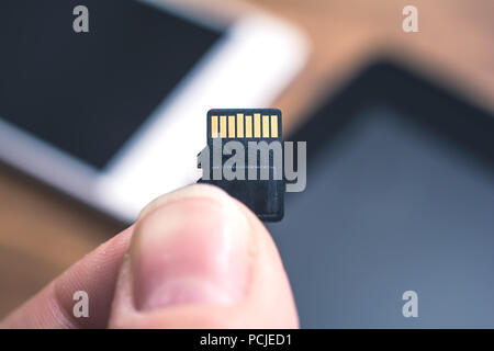 Male Fingers Holding A Micro SD Card With Tablet And Smartphone In Background, Extent Disk Space Concept Stock Photo