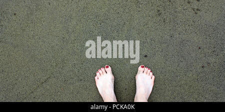 A woman's feet with red painted toenails on black and green sand at the Papakolea Beach, also known as the Green Sand Beach in Hawaii, USA Stock Photo