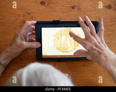 An elderly woman playing mahjong on a tablet that's resting a wooden table. Only her hands and top of her head are showing. Stock Photo