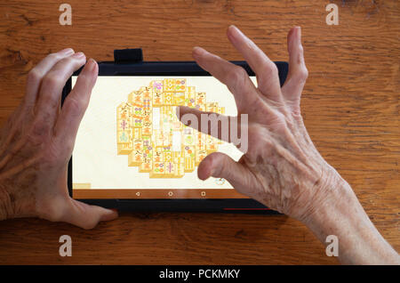 An elderly woman playing mahjongg on a tablet that's resting a wooden table. Only her hands are showing. Stock Photo