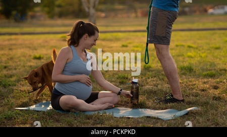 Young healthy pregnant woman with two red dogs doing yoga exercises in nature outdoors on green grass on fitness mat at sunset. Happy pregnancy and motherhood concept Stock Photo